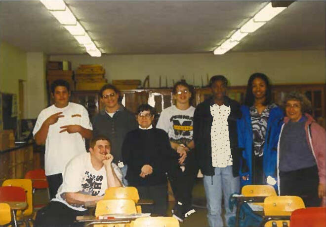 Students in classroom sitting and standing around desks smiling at camera